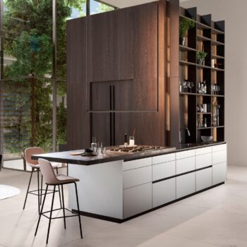 Leicht Küchen Contino E12 - Moderne Küche mit großer Insel, eingebautem Herd, Spüle, zwei Barhockern, offenen Regalen und hohen Holzschränken, mit großen Fenstern, die den Blick ins Grüne freigeben.