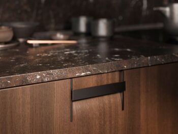 Leicht Küchen Griffkonzept Q - Eine Nahaufnahme einer Arbeitsplatte aus dunklem Marmor von Leicht Küchen mit Holzschränken darunter, mit dem eleganten Griffkonzept Q, schwarzen Metallgriffen und Kochgeschirr im Hintergrund.