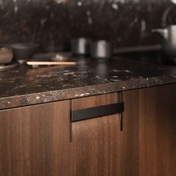 Leicht Küchen Griffkonzept Q - Eine Nahaufnahme einer Arbeitsplatte aus dunklem Marmor von Leicht Küchen mit Holzschränken darunter, mit dem eleganten Griffkonzept Q, schwarzen Metallgriffen und Kochgeschirr im Hintergrund.