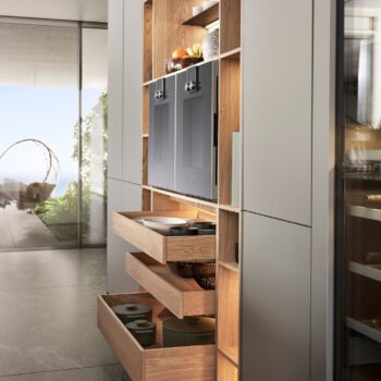 Leicht Küchen KYOTO - Moderne Leicht Küchen Küche mit offenen Holzregalen und KYOTO-Schubladen, in denen Kochgeschirr, Geschirr und Geräte neben einem Weinkühler aufbewahrt werden; ein großes Fenster bietet einen einladenden Blick nach draußen.