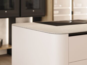 Leicht Küchen Rundwangen - Nahaufnahme einer modernen Leicht Küchen Insel im Rundwangen-Design mit heller Arbeitsplatte und eingebautem Kochfeld; im Hintergrund sind Wandöfen und Regale zu sehen.