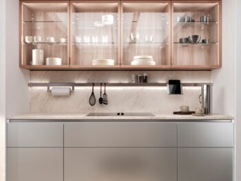 Leicht Küchen in der Trendfarbe Beige - Moderne Küche mit Holzschränken mit Glasfronten, in denen Geschirr und Gläser ausgestellt sind, Marmorsockel, Unterschrankbeleuchtung und minimalistische Arbeitsplatte aus Metall und Stein.