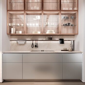 Leicht Küchen in der Trendfarbe Beige - Moderne Küche mit Holzschränken mit Glasfronten, in denen Geschirr und Gläser ausgestellt sind, Marmorsockel, Unterschrankbeleuchtung und minimalistische Arbeitsplatte aus Metall und Stein.