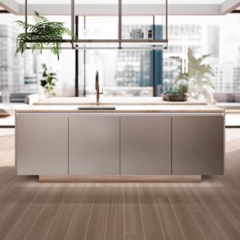 Leicht Küchen in der Trendfarbe Beige - Moderne Kücheninsel mit Metallic-Finish, eingebauter Spüle und Ablagefläche über dem Kopf in einem hellen Raum mit großen Fenstern und Stadtblick.