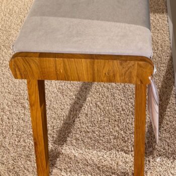 Ein Holzhocker im Retro-Massivholzbank-Stil mit einem hellgrauen gepolsterten Sitz sitzt auf einem beigen Teppich. An einem der Beine ist ein weißes Schildchen befestigt.