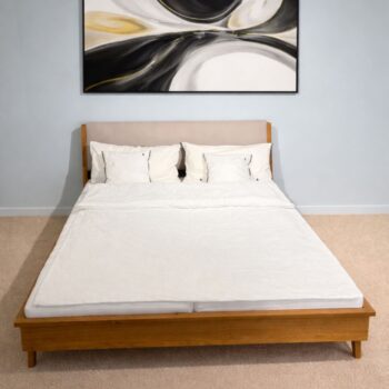 Ein ordentlich gemachtes Retro-Massivholzbett mit weißem Bettzeug und drei Kissen steht auf einem beigen Teppich, über dem Kopfteil hängt abstrakte Kunst an einer hellen Wand.
