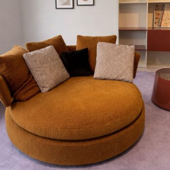 Ein großes, rundes, orange-braunes Maxalto Amoenus Sofa mit vier quadratischen Kissen steht auf einem hellvioletten Teppich in einem modernen Wohnzimmer und strahlt Luxus und Komfort aus.