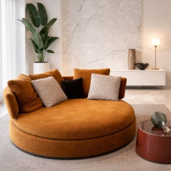 Ein großes rundes Maxalto Amoenus Sofa mit verschiedenen Kissen steht auf einem Teppich neben einem kleinen roten Tisch in einem modernen Wohnzimmer mit Marmorwand und Zimmerpflanze.