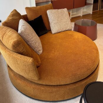 Das runde, rostfarbene Maxalto Amoenus Sofa mit drei Wurfkissen steht auf einem hellen Teppich neben einem braunen Beistelltisch in einem modernen Wohnzimmer.