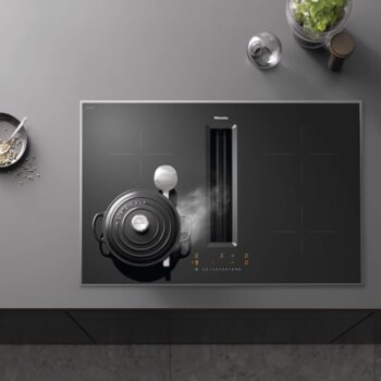Auf der grauen Arbeitsplatte steht ein mattschwarzes Miele-Kochfeld mit Dampftopf und Utensilien, daneben eine Schüssel mit Grünzeug und ein kleines Glas.