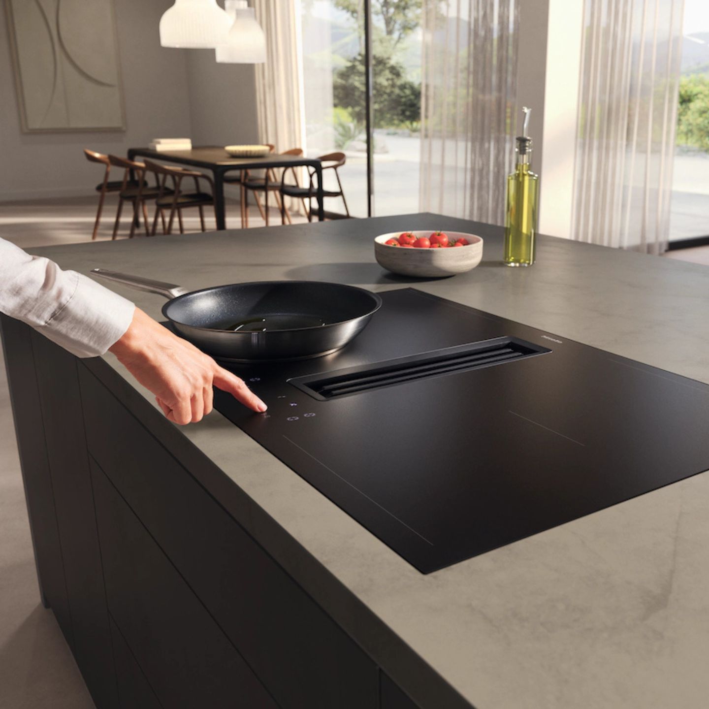 Eine Person berührt das Bedienfeld eines mattschwarzen Miele-Kochfelds mit Induktionskochfeld, auf dem eine Pfanne steht, die in einer Kücheninsel platziert ist; daneben stehen Öl und eine Schüssel mit Tomaten.