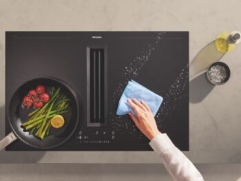 Eine Person wischt mit einem blauen Tuch verschüttetes Salz von einem mattschwarzen Miele-Kochfeld; daneben stehen eine Bratpfanne mit Gemüse und eine Flasche Öl.