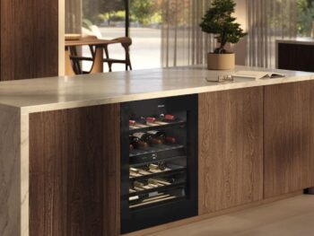 Moderne Kücheninsel mit eingebautem Miele Weinkühlschrank KWTUS 7196 E, eleganter Marmorarbeitsplatte, Holzschränken und einer kleinen Topfpflanze darauf. Große Fenster im Hintergrund füllen den Raum mit natürlichem Licht.