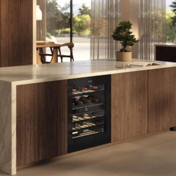 Moderne Kücheninsel mit eingebautem Miele Weinkühlschrank KWTUS 7196 E, eleganter Marmorarbeitsplatte, Holzschränken und einer kleinen Topfpflanze darauf. Große Fenster im Hintergrund füllen den Raum mit natürlichem Licht.