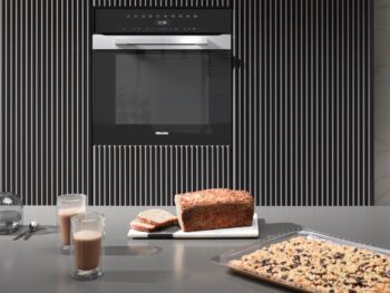 Ein moderner Miele-Backofen H 7464 BP ist in eine gestreifte Wand eingelassen; auf dem Tresen stehen zwei Gläser Schokomilch, ein Brot und ein Tablett mit gebackenem Dessert mit Obst oder Nüssen.