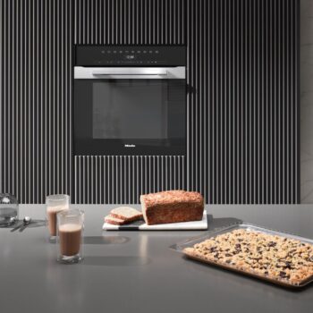 Ein moderner Miele-Backofen H 7464 BP ist in eine gestreifte Wand eingelassen; auf dem Tresen stehen zwei Gläser Schokomilch, ein Brot und ein Tablett mit gebackenem Dessert mit Obst oder Nüssen.