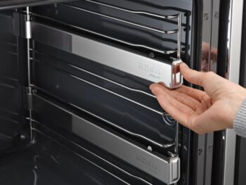 Eine Hand verstellt ein Metallgestell im Inneren eines modernen Miele-Backofens H 7464 BP mit schlichtem grauem Innenraum.