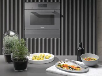 Eine moderne Küche mit einem Einbaubackofen, Miele Dampfgarer DGM 7440, Kräutertöpfen, einem Teller mit Zitronen, einem ganzen Fisch mit Gemüse, einer Salatschüssel und einer Flasche auf einer grauen Arbeitsplatte.