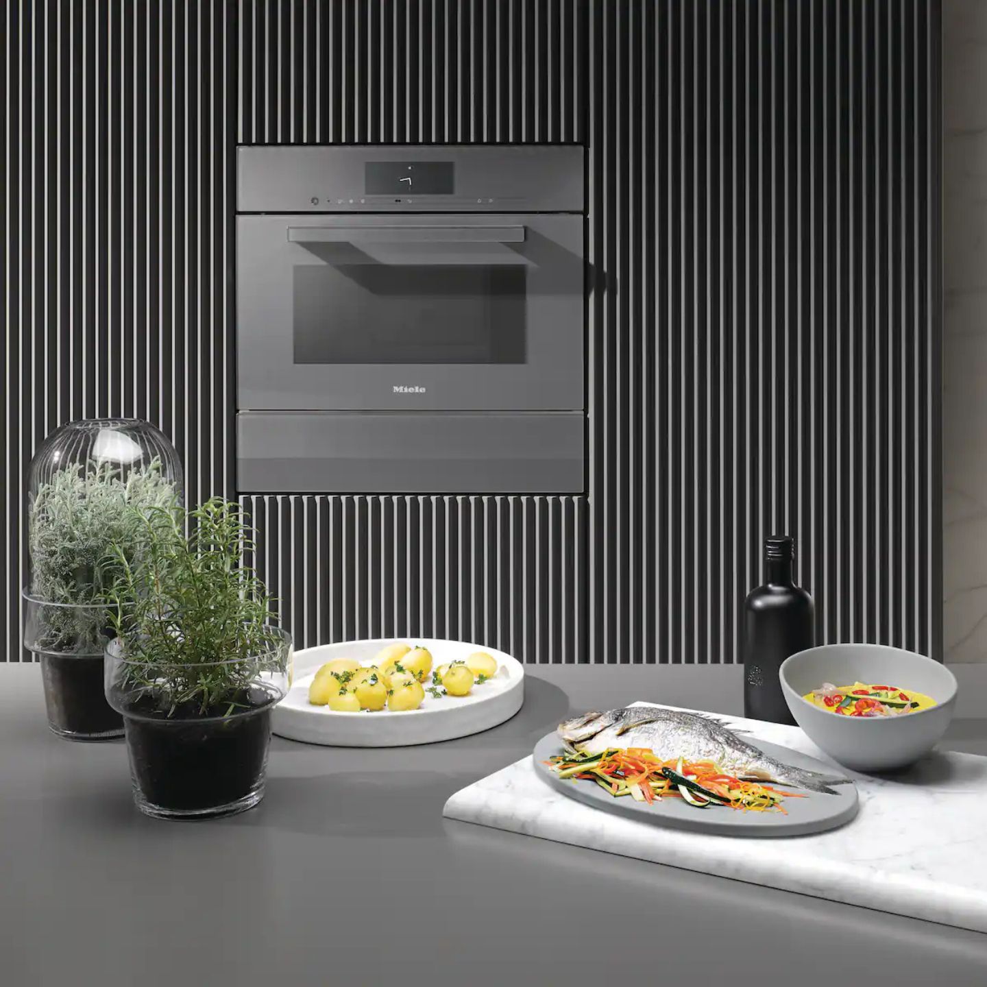 Eine moderne Küche mit einem Einbaubackofen, Miele Dampfgarer DGM 7440, Kräutertöpfen, einem Teller mit Zitronen, einem ganzen Fisch mit Gemüse, einer Salatschüssel und einer Flasche auf einer grauen Arbeitsplatte.