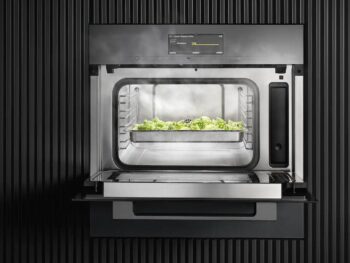 Der Miele-Dampfgarer DGM 7440 zeigt bei geöffneter Tür eine Schale mit gedämpftem Gemüse vor einem dunklen, vertikalen Hintergrund. Das vielseitige Gerät verfügt auch über eine integrierte Mikrowelle.