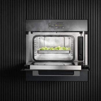 Der Miele-Dampfgarer DGM 7440 zeigt bei geöffneter Tür eine Schale mit gedämpftem Gemüse vor einem dunklen, vertikalen Hintergrund. Das vielseitige Gerät verfügt auch über eine integrierte Mikrowelle.