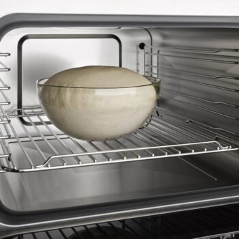 Eine Glasschüssel mit aufgegangenem Teig steht auf einem Gitterrost in einem leeren Edelstahl-Backofen, bereit zum Backen - perfekt für den Einsatz im Miele-Dampfgarer oder zum Aufwärmen in der Mikrowelle.