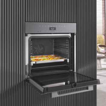 Ein moderner Einbaubackofen mit offener Tür, in der eine Schale mit verschiedenen Gemüsesorten zu sehen ist, vor einer vertikalen, schwarz getäfelten Wand - die perfekte Ergänzung zu Ihrem Miele Dampfgarer oder Ihrer Mikrowelle für ein nahtloses Küchenerlebnis.