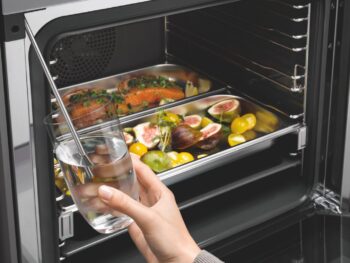 Eine Hand hält ein Glas mit Wasser, in das eine Sonde eingeführt ist, vor einem offenen Backofen, in dem sich Bleche mit Fisch, Feigen, Tomaten und Kräutern befinden - die perfekte Szene, um die Präzision eines Miele-Dampfgarers zu testen.