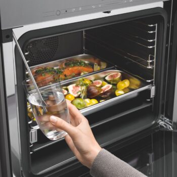 Eine Hand hält ein Glas mit Wasser, in das eine Sonde eingeführt ist, vor einem offenen Backofen, in dem sich Bleche mit Fisch, Feigen, Tomaten und Kräutern befinden - die perfekte Szene, um die Präzision eines Miele-Dampfgarers zu testen.