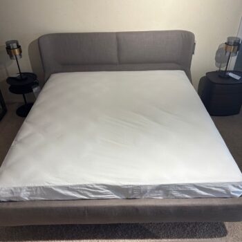 Ein sauber verarbeitetes Polsterbett mit weißem Laken und grau gepolstertem Kopfteil, flankiert von zwei dunklen Nachttischen mit Lampen auf einem Teppichboden, bringt den raffinierten Stil der Fulham-Kollektion von Molteni&C ins Schlafzimmer.