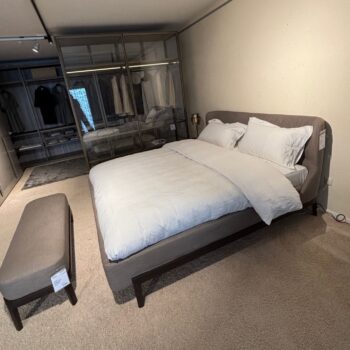 Modernes Schlafzimmer mit einem Molteni&C Polsterbett mit weißer Bettwäsche, einem gepolsterten Fulham-Kopfteil, einer Bank am Fußende und einem verglasten Kleiderschrank mit aufgehängter Kleidung im Hintergrund.