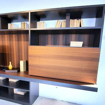 Ein modernes Bücherregal mit Holzregalböden, mehreren aufrecht stehenden und gestapelten Büchern, einer kleinen gelben Vase und einer dekorativen Schachtel auf dem unteren Regalboden – all dies spiegelt das schlichte Design wider, das vom charakteristischen Stil von Molteni inspiriert ist und an deren ikonische „505 Anbauwand“ erinnert.