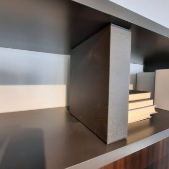 Eine Nahaufnahme eines modernen Regals – Teil des eleganten Molteni 505 Anbauwand-Systems – zeigt eine große schwarze Buchstütze neben einem Stapel Bücher mit hellen Einbänden. Das Regal hat eine dunkle, matte Oberfläche.