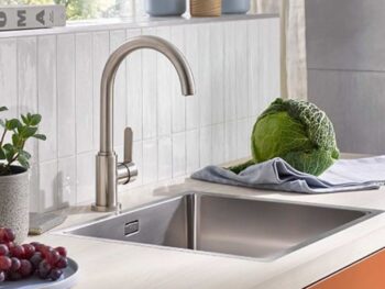 Eine moderne Küchenspüle mit einem Wasserhahn aus rostfreiem Stahl, ein Kohlkopf auf einem Tuch, Weintrauben, eine Topfpflanze und Keramikbehälter auf der Arbeitsplatte.