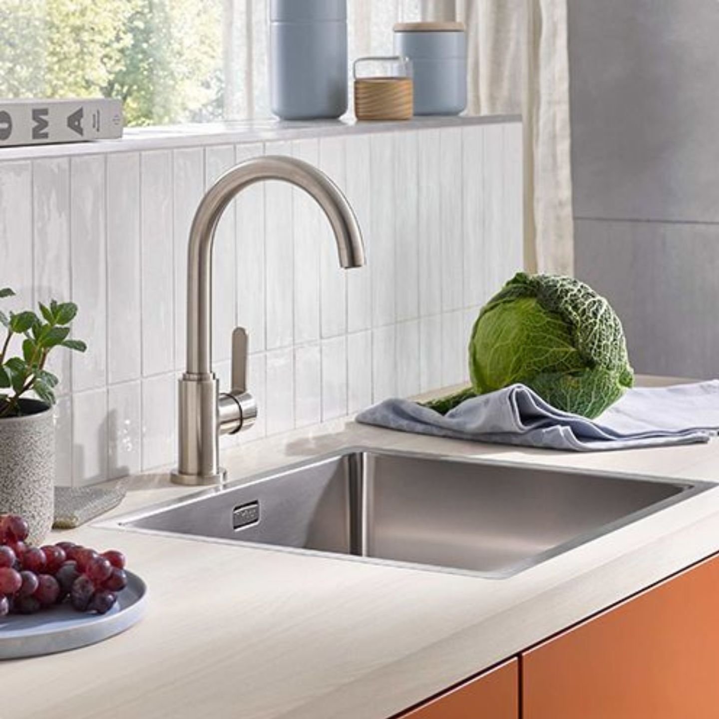 Eine moderne Küchenspüle mit einem Wasserhahn aus rostfreiem Stahl, ein Kohlkopf auf einem Tuch, Weintrauben, eine Topfpflanze und Keramikbehälter auf der Arbeitsplatte. Eine moderne Küchenspüle mit einem Wasserhahn aus rostfreiem Stahl, ein Kohlkopf auf einem Tuch, Weintrauben, eine Topfpflanze und Keramikbehälter auf der Arbeitsplatte.