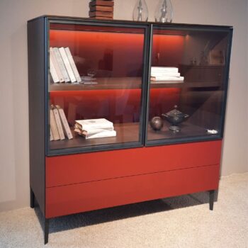 Das 505 UP Highboard von Molteni&C ist ein moderner Schrank mit Glasfront und roten unteren Schubladen, der Bücher und Dekorationsgegenstände auf zwei Regalen präsentiert, die von einem warmen orangefarbenen Licht beleuchtet werden.