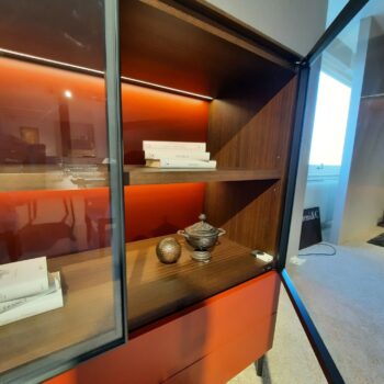 Ein Molteni&C 505 UP Highboard mit roten Holzregalen präsentiert Bücher, einen dekorativen silbernen Behälter und eine strukturierte Kugel. Die Glastür des Schranks ist geöffnet.