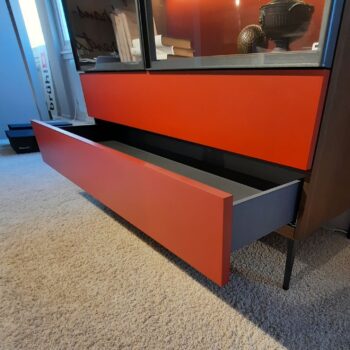Ein modernes Molteni&C 505 UP Highboard mit einer teilweise geöffneten roten Schublade, die den Blick auf ein leeres Inneres freigibt, steht auf einem beigen Teppichboden.