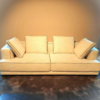 Ein beige gepolstertes Molteni&C Augusto-Sofa mit vier losen Kissen steht vor einer schlichten hellbraunen Wand auf einem dunklen Teppichboden.