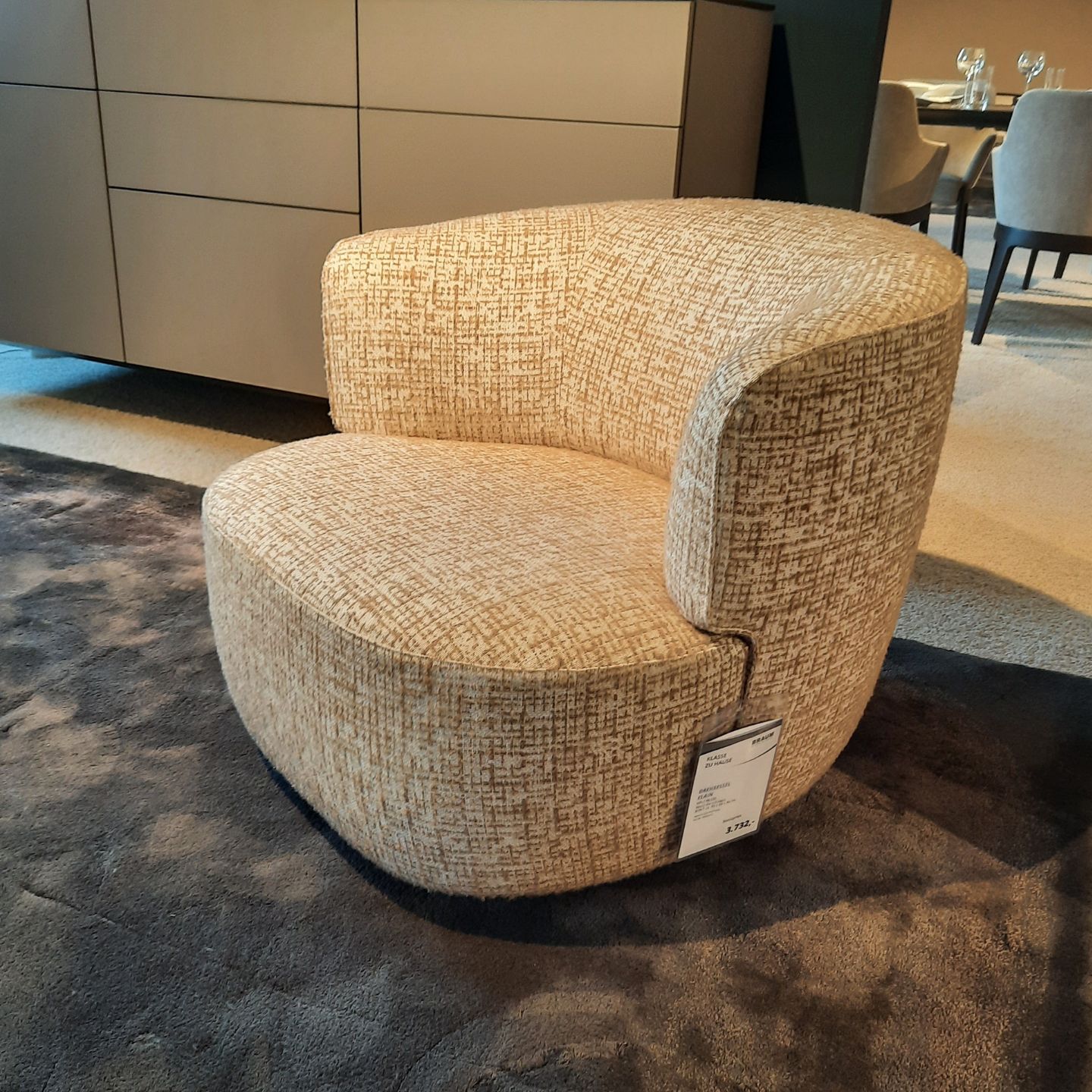 Ein moderner, beige gepolsterter Elain-Drehsessel von Molteni&C mit geschwungener Rückenlehne steht auf einem dunklen Teppich in einem Ausstellungsraum, an seinem Fuß ist ein Preisschild befestigt.