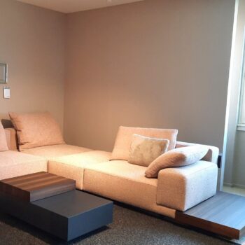 Ein minimalistisches Wohnzimmer verfügt über ein Marteen Polstereckgarnitur-Sofa von Molteni&C mit Kissen, einen braunen Couchtisch mit Stufen und Wände in neutralen Farben mit einem Fenster, das natürliches Licht hereinlässt.