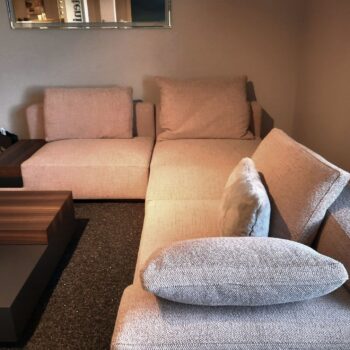 Ein modernes beiges Marteen-Sofa mit zwei großen und einem kleinen Kissen steht neben einem dunklen Couchtisch aus Holz in einem mit Teppich ausgelegten Wohnzimmer.