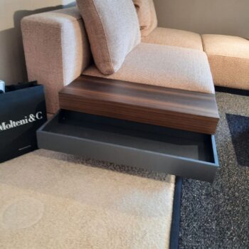 Eine Schublade mit Holzplatte eines modernen Polstereckgarnitur-Sofas öffnet sich und gibt den Blick auf ein leeres, graues Inneres frei. Auf dem beigefarbenen Teppich daneben liegt eine schwarze Einkaufstasche, die einen Hauch von Eleganz verleiht und an Marteen von Molteni&C erinnert.