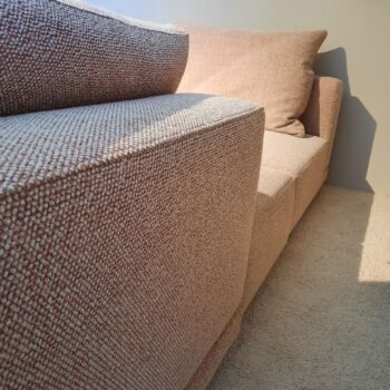 Nahaufnahme einer strukturierten beigen Molteni&C Marteen Polstereckgarnitur mit passendem Kissen auf einem hellen Teppich, vor einer schlichten Wand im natürlichen Tageslicht platziert.