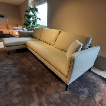 Ein beiges L-förmiges Sofa „Paul“ von Molteni&C mit drei Kissen steht auf einem braunen Teppich in einem modernen Wohnzimmer mit neutralen Wänden und einer Topfpflanze in der Nähe.