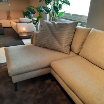 Ein beiges Molteni&C-Sofa mit strukturiertem Kissen steht in einem Wohnzimmer mit einer Pflanze, einem Fenster und weiteren Sitzgelegenheiten im Hintergrund.