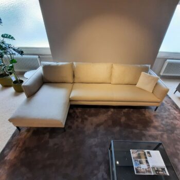 Ein beiges Molteni-Sofaelement mit einer Chaiselongue auf der linken Seite, platziert auf einem braunen Teppich neben einem Couchtisch aus Glas mit Zeitschriften, neben Topfpflanzen und großen Milchglasfenstern.