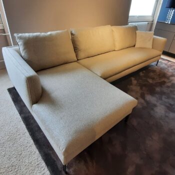 Ein beiges Molteni&C-Sofa in L-Form mit drei Kissen steht auf einem dunklen Teppich in einem modernen Wohnzimmer mit neutralen Wänden.