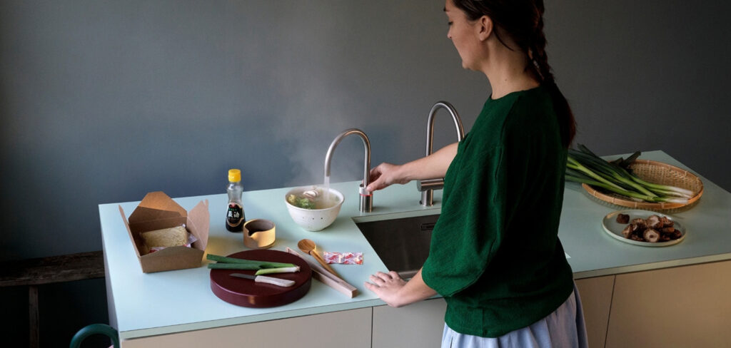 Eine Frau steht an einer Küchentheke neben einer dampfenden Suppenschüssel; daneben sind ein Quooker-Wasserhahn sowie Zutaten wie Sojasauce, Frühlingszwiebeln und Pilze zu sehen.