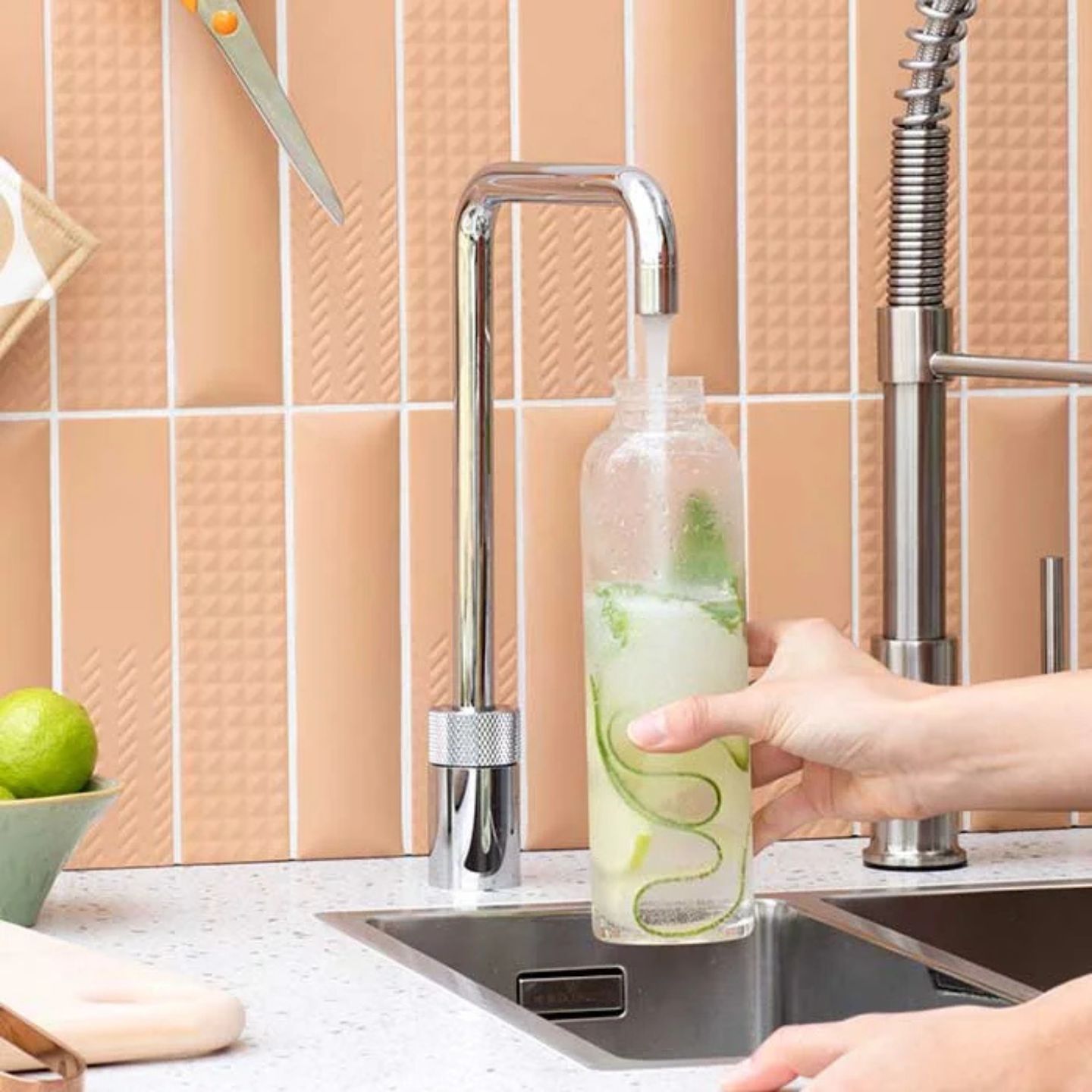 Eine Hand hält eine durchsichtige Flasche mit Wasser, Limette und Minze unter einem laufenden Küchenwasserhahn neben einem Waschbecken mit beigefarbener Kacheloberfläche.